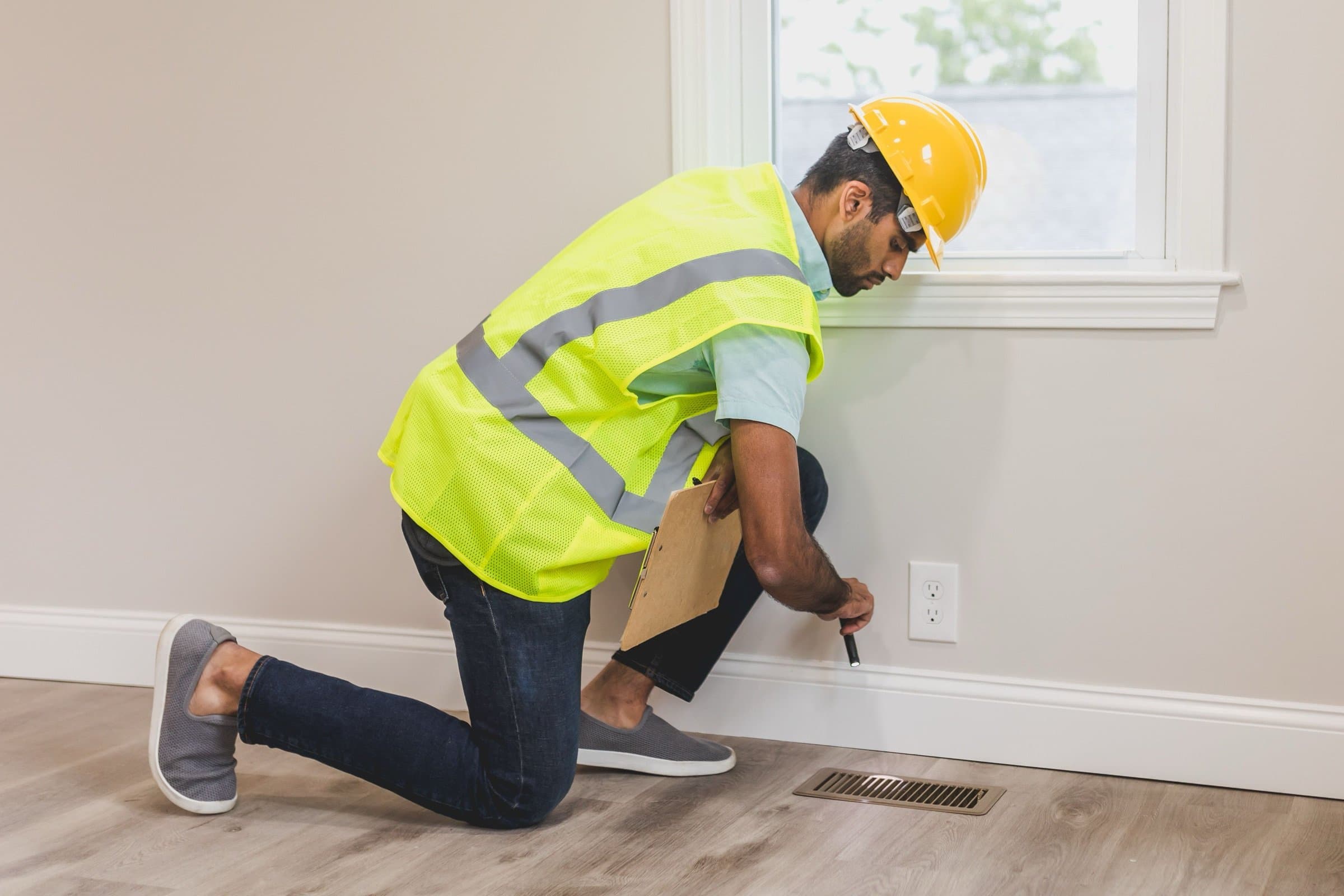 An inspector in hi-vis kneeling at a baseboard vent with a clipboard