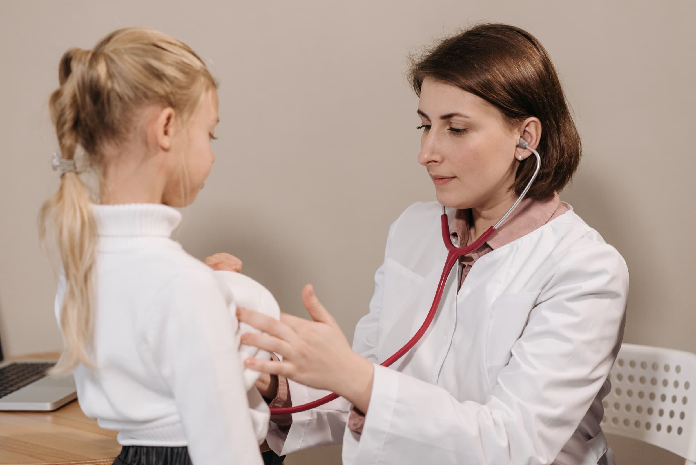 A pediatrician in a white lab coat listening to a young child's breathing with a stethoscope